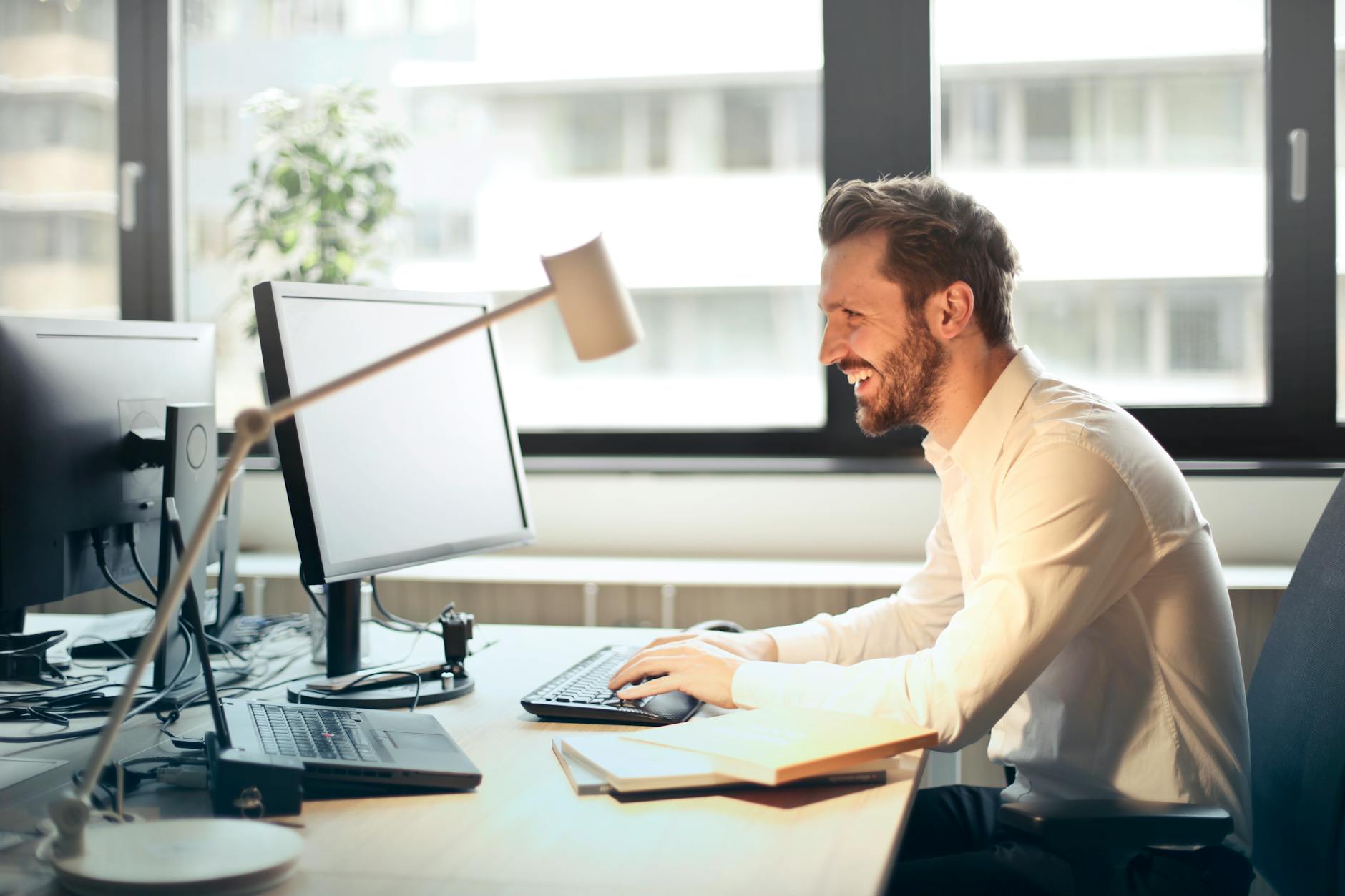 man in white dress shirt sitting on black rolling chair while facing black computer set and smiling - retirement plan participants